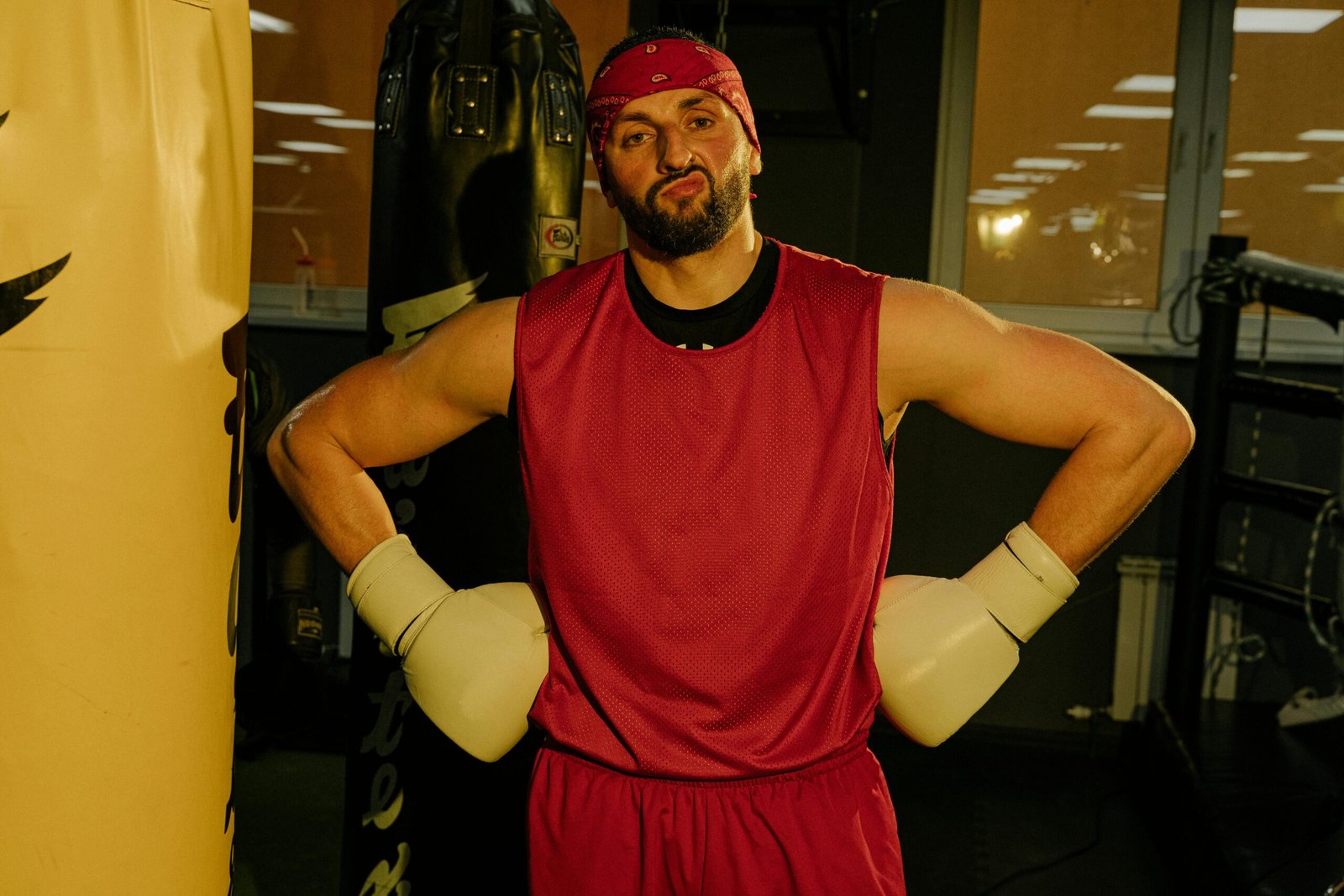 A muscular man with a beard flexes confidently in a boxing gym.